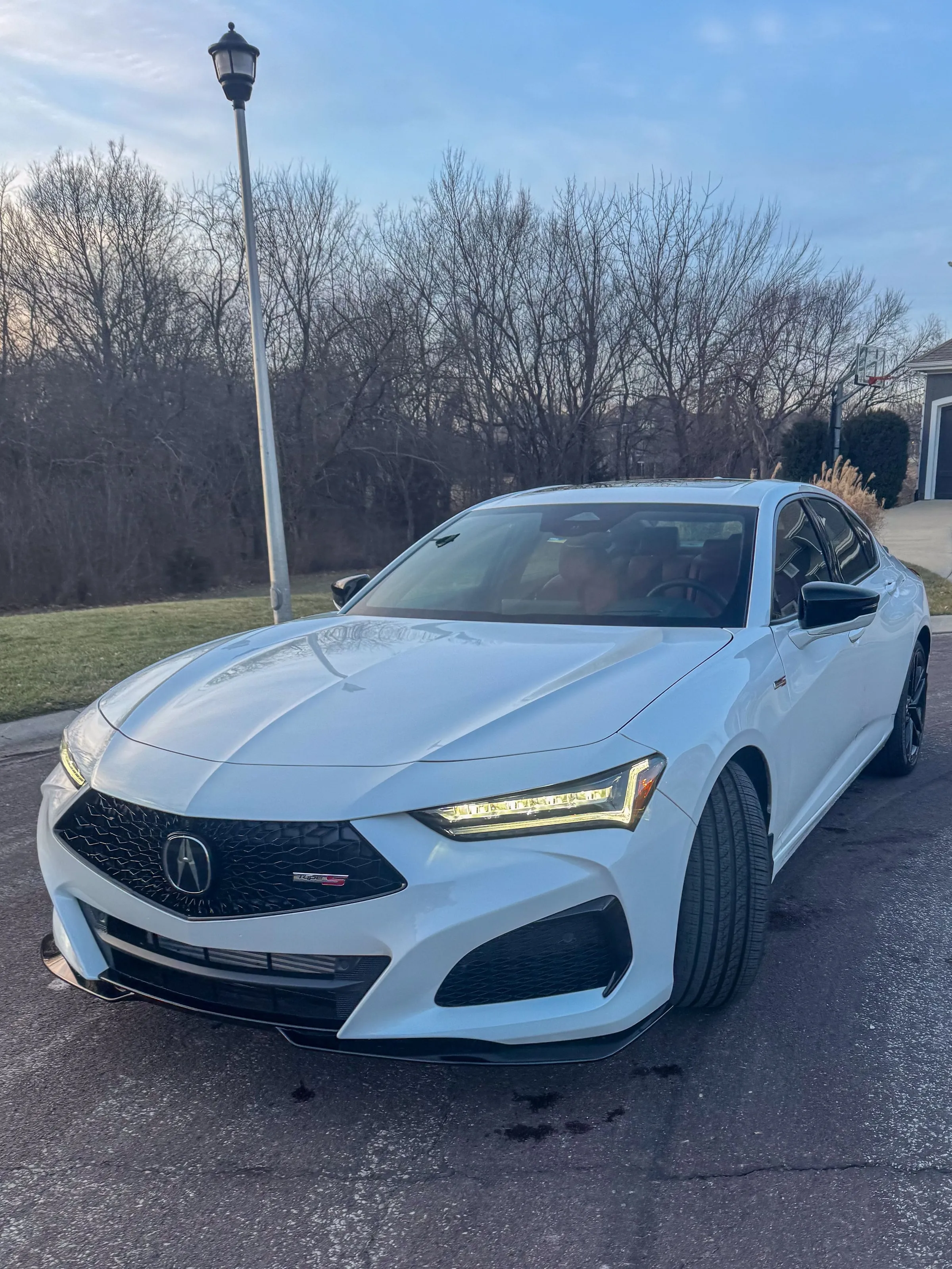 White Acura TLX Type S on a residential street at golden hour, headlights lit and paint reflecting bare trees