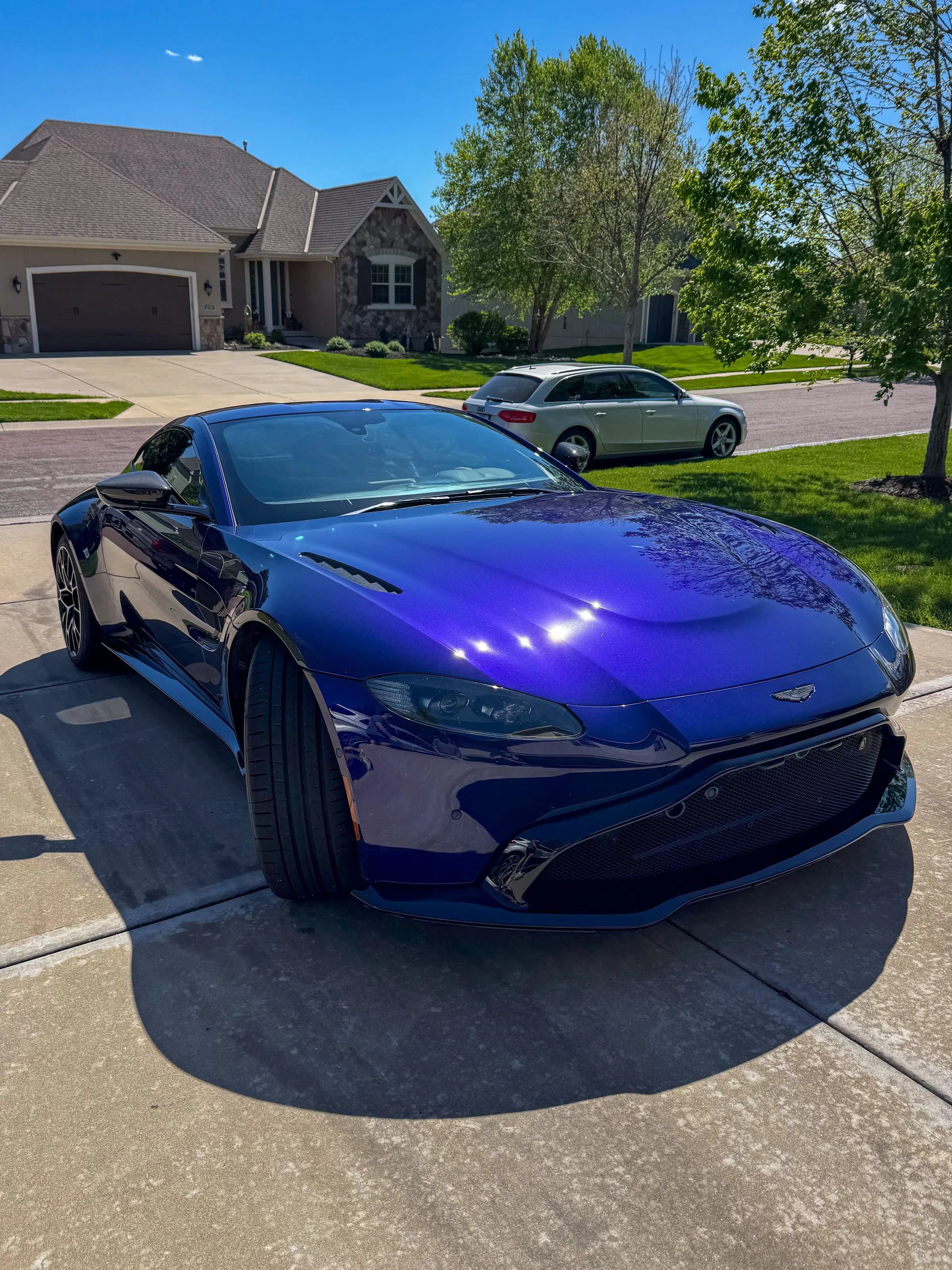 Deep blue Aston Martin Vantage on a concrete driveway in full sun, paint reflecting the sky and the treeline