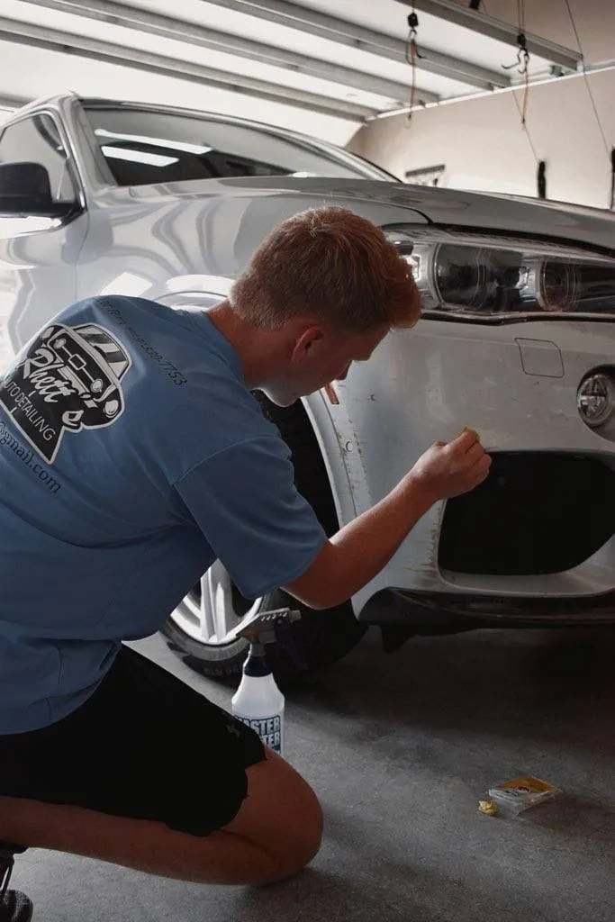 Rhett kneeling beside a white BMW X5, working a product into the lower front bumper by hand
