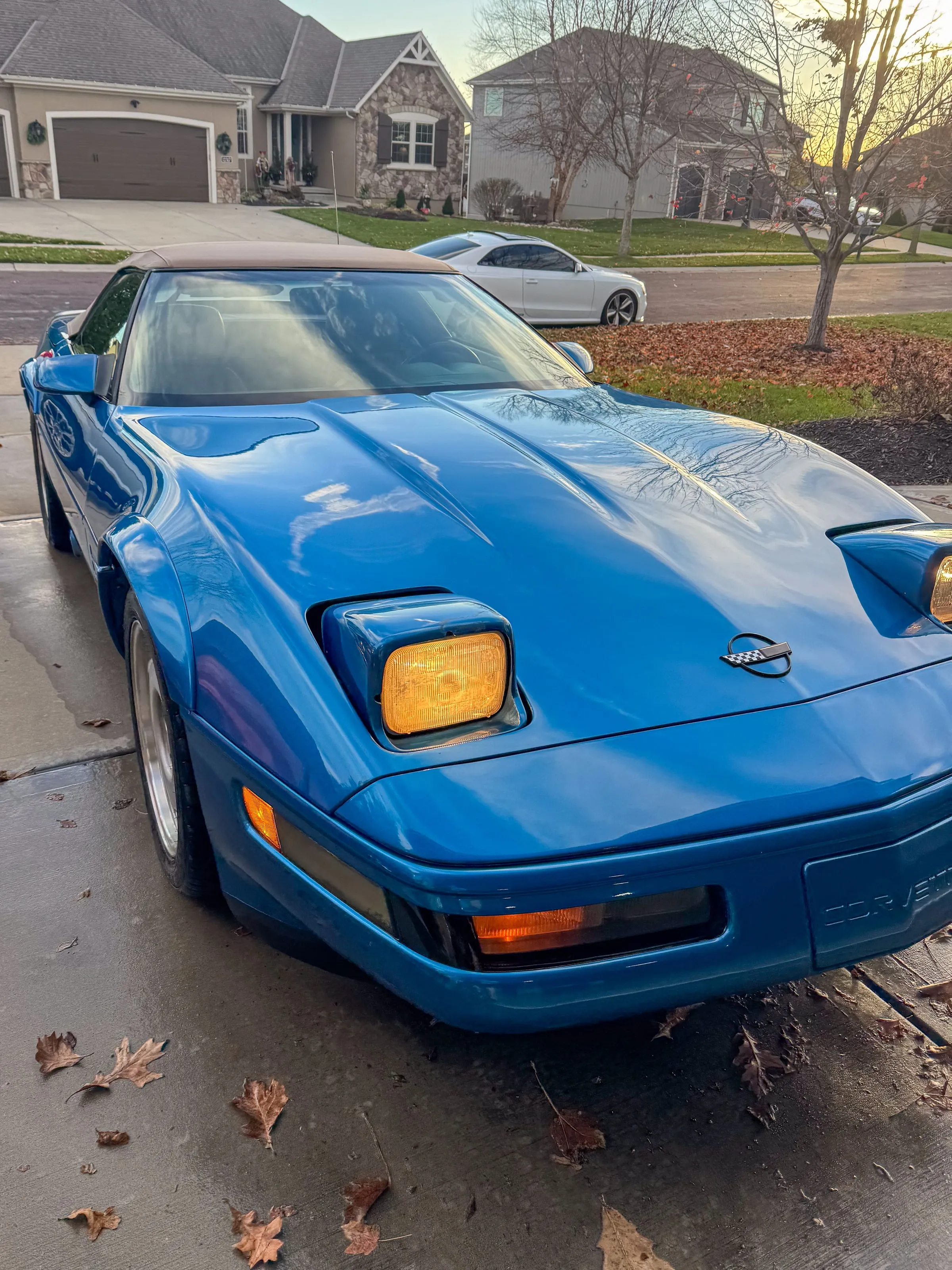 Blue Corvette convertible with a tan soft top on a driveway at dusk, one pop-up headlight raised