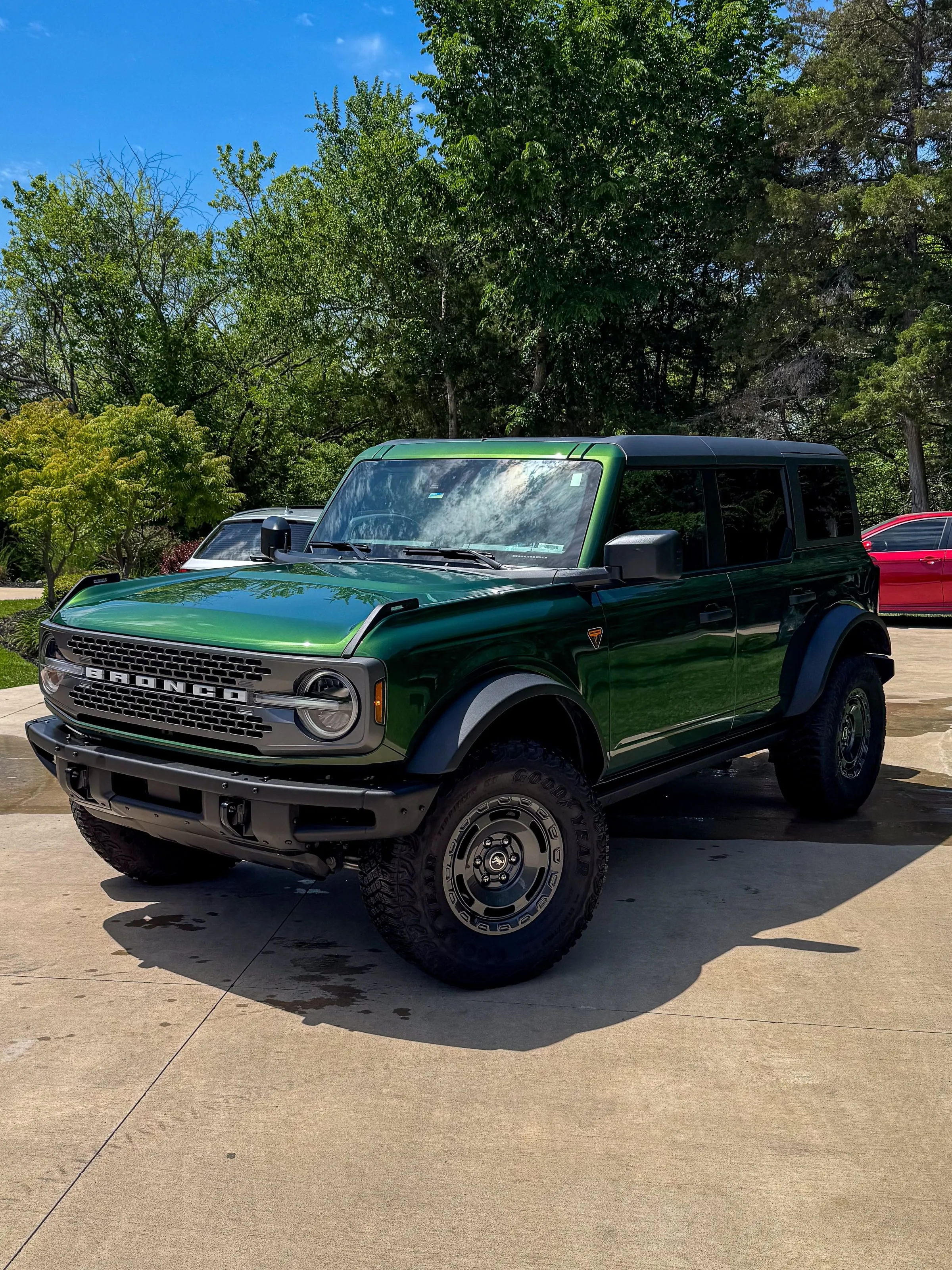 Green four-door Ford Bronco on a driveway with tires dressed and paint cleaned, woods behind it