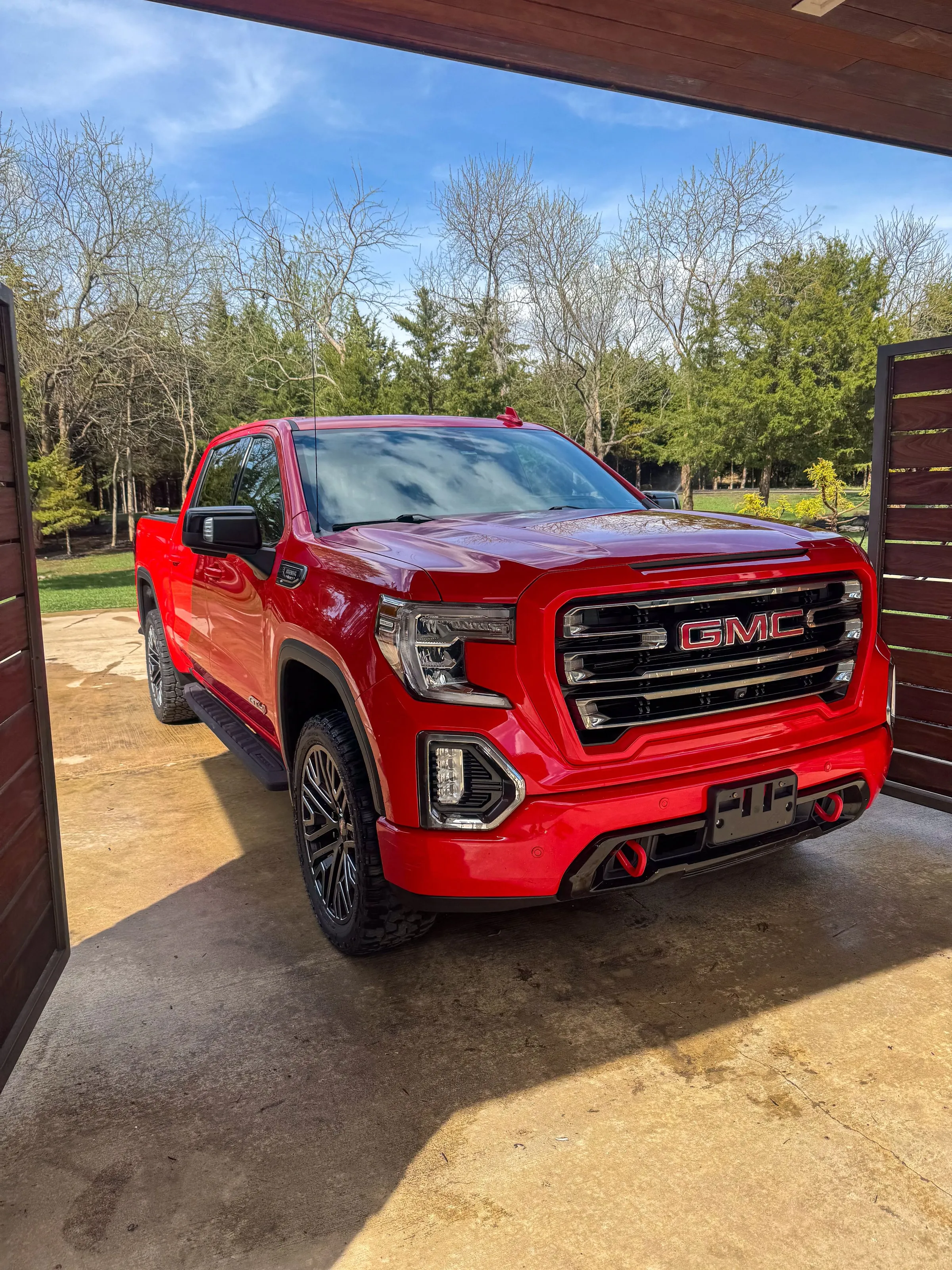 Red GMC Sierra crew cab parked under a covered slab, grille and wheels cleaned, trees behind it