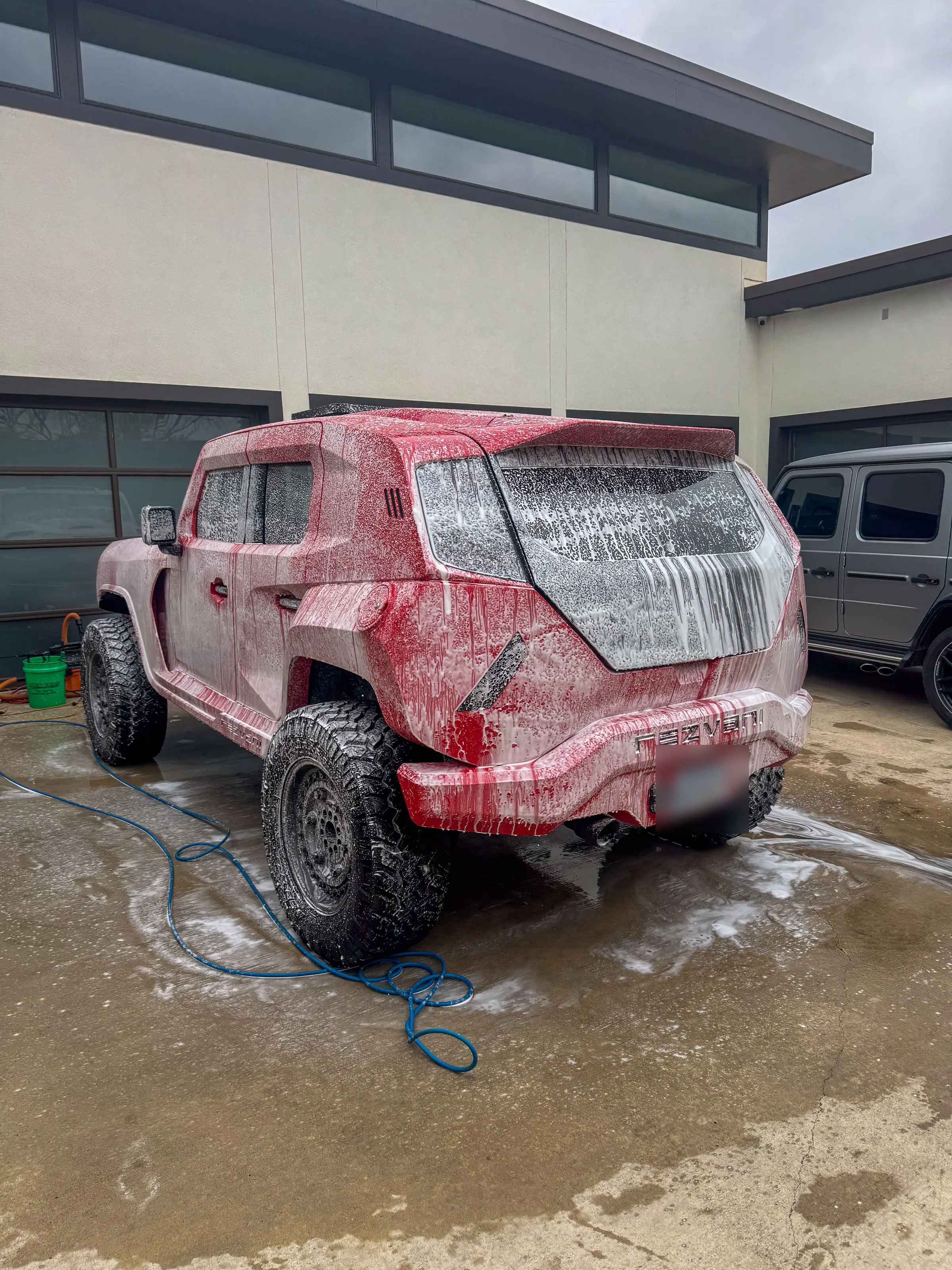 Red Rezvani Tank covered in wash foam on a driveway, hose run out and the rinse already running off