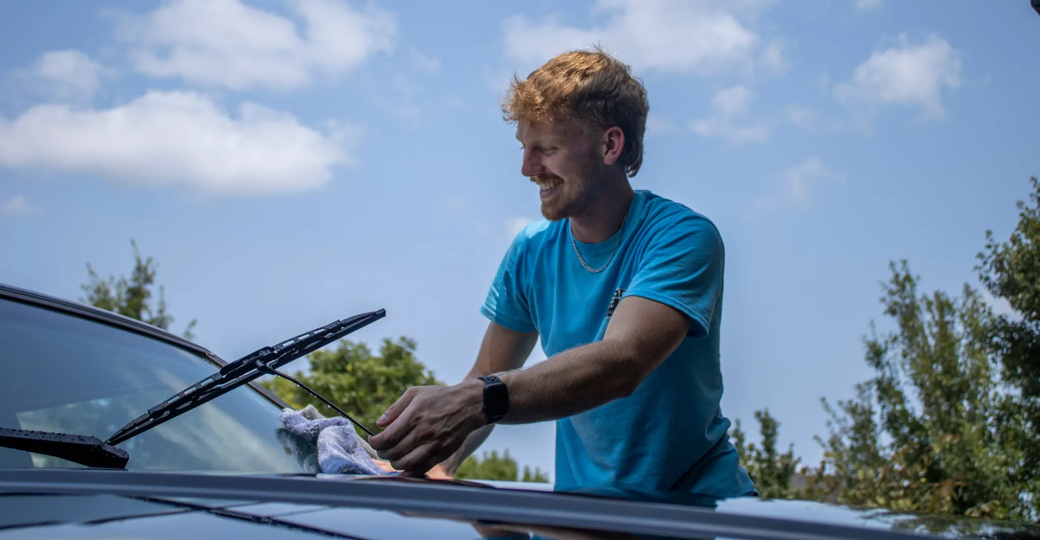 Rhett smiling while wiping down the base of a vehicle's windshield with a microfiber towel