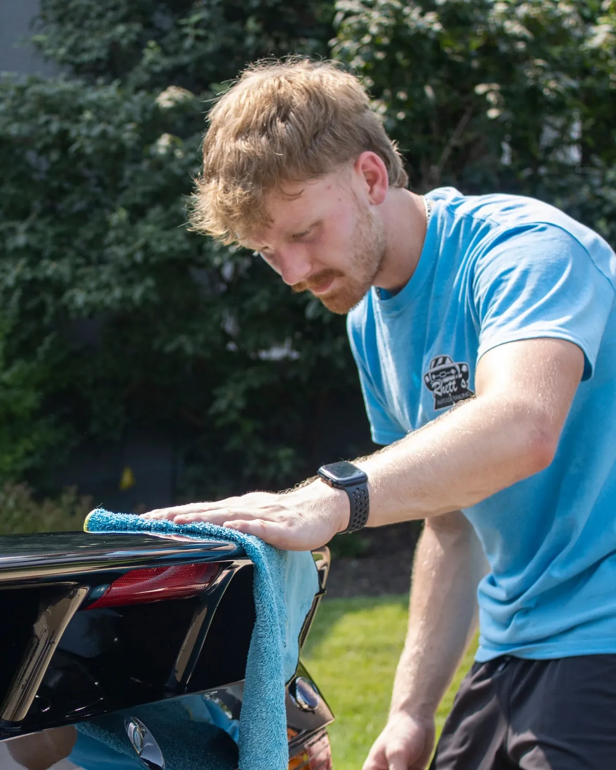 Rhett leaning over the rear decklid of a black Mustang, drying it with a microfiber towel