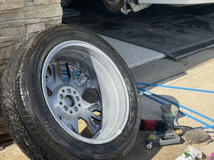 A white SUV up on ramps with a wheel removed, the bare hub and brake caliper visible, wash gear on the driveway